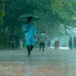 A man in blue walking under heavy rain with umbrella on a tree-lined path.